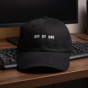 May include: A black baseball cap featuring the white embroidered text OFF BY ONE centered on the front panel. The cap has a structured crown, a curved brim, and a button at the top center. The fabric appears to be a durable cotton twill material.