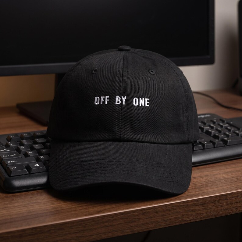 May include: A black baseball cap featuring the white embroidered text OFF BY ONE centered on the front panel. The cap has a structured crown, a curved brim, and a button at the top center. The fabric appears to be a durable cotton twill material.