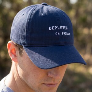 Pode incluir: Bon&eacute; de beisebol azul marinho com o texto "DEPLOYED ON FRIDAY" em branco. O bon&eacute; tem uma aba curva e um bot&atilde;o na parte superior. O chap&eacute;u &eacute; feito de um material macio e dur&aacute;vel.