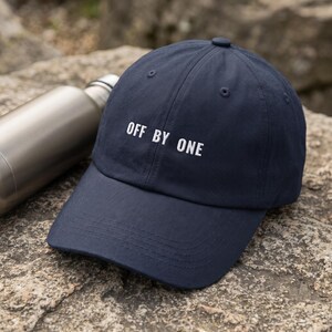 May include: A navy blue baseball cap featuring the white embroidered text OFF BY ONE centered on the front panel. The cap has a structured crown, a curved brim, and stitched eyelets, presenting a clean and simple design for casual headwear.