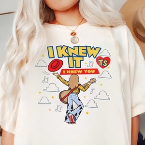 Puede incluir: Una camiseta color hueso con un estampado gr&aacute;fico de una persona de pelo largo tocando la guitarra, vistiendo una camisa de cuadros amarilla y pantalones acampanados con estampado de vaca. El texto dice I KNEW IT I KNEW YOU, con nubes y estrellas.