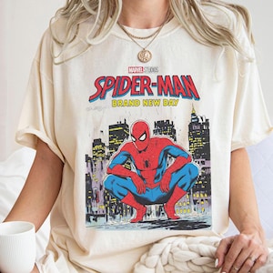 K&ouml;nnte beinhalten: Cremefarbenes T-Shirt mit einer Grafik von Spider-Man in einer hockenden Pose vor einer Stadtkulisse. Der Text "MARVEL STUDIOS SPIDER-MAN BRAND NEW DAY" ist in Rot und Blau &uuml;ber dem Bild gedruckt.