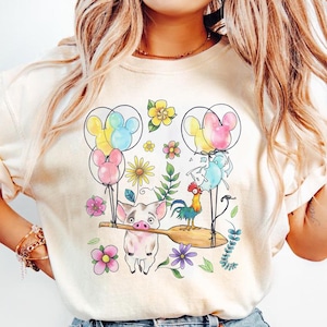 K&ouml;nnte beinhalten: Cremefarbenes T-Shirt mit einem farbenfrohen Aufdruck eines Schweins, das ein Holzpaddel mit einem Hahn, Luftballons und Blumen tr&auml;gt. Die Ballons sind wie Mickey Mouse-K&ouml;pfe geformt. Das Design umfasst verschiedene Blumen und Bl&auml;tter.
