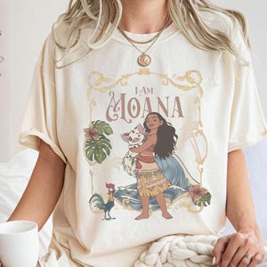 May include: Cream-colored t-shirt featuring a graphic of Moana holding a pig, with the text "I AM MOANA" in a decorative frame. The design includes tropical flowers and a rooster. The shirt has short sleeves.