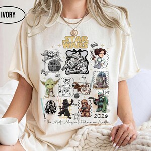 May include: Ivory-colored t-shirt featuring a Star Wars graphic with various characters and elements from the franchise. The design includes illustrations of characters like Yoda, Darth Vader, and Chewbacca, with the text "Star Wars" and "2026".