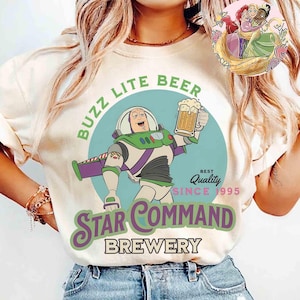 Könnte beinhalten: Cremefarbenes T-Shirt mit einem Cartoon-Design von Buzz Lightyear, der einen Bierkrug hält. Der Aufdruck enthält den Text "BUZZ LITE BEER" und "STAR COMMAND BREWERY" in Grün und Lila. Das Shirt hat auch die Worte "Best Quality Since 1995".