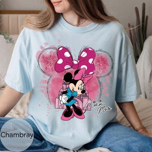 Op de afbeelding: Lichtblauw T-shirt met een Minnie Mouse-afbeelding. Het ontwerp bevat een roze strik met stippen, een kasteel en de woorden "Minnie Mouse" in cursief. Het shirt is gemaakt van een zacht materiaal en is geschikt voor vrijetijdskleding.