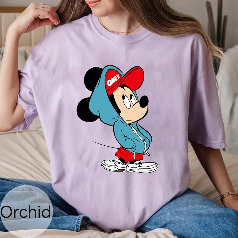 May include: A light purple t-shirt featuring a cartoon Mickey Mouse wearing a blue hoodie and a red baseball cap with the word "OBEY". Mickey is also wearing red shorts and white sneakers. The shirt has a relaxed fit.
