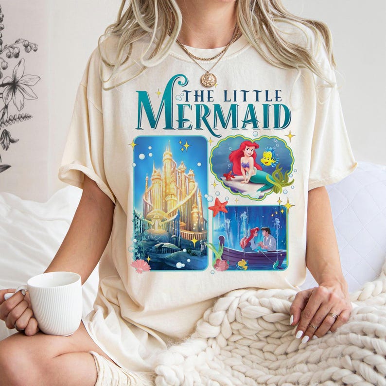 May include: Cream-colored t-shirt featuring the text "THE LITTLE MERMAID" in teal script. The shirt has three graphic panels: a golden castle, Ariel and Flounder, and Ariel and Eric in a boat. The shirt has short sleeves.