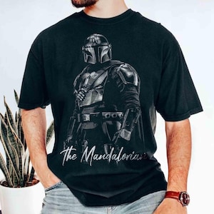 May include: Black t-shirt featuring a detailed illustration of The Mandalorian character in armor. The image is in grayscale with the text "The Mandalorian" in a cursive font at the bottom. The shirt is a crew neck.