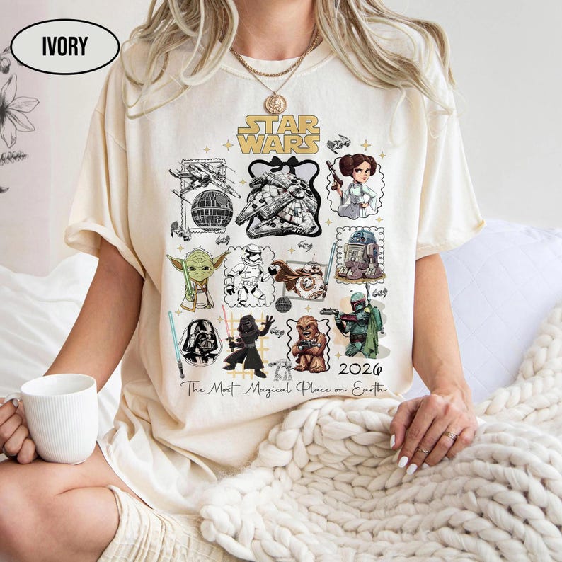 May include: Ivory-colored t-shirt featuring a Star Wars graphic with various characters and elements from the franchise. The design includes illustrations of characters like Yoda, Darth Vader, and Chewbacca, with the text "Star Wars" and "2026".
