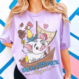May include: A light purple t-shirt featuring a cartoon illustration of a pig in a boat with a rooster, ice cream, and donuts. The text "BOAT SNACK Upgrade" is printed in blue and gold script. The shirt has short sleeves.
