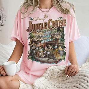 May include: A light pink t-shirt featuring a Disney Jungle Cruise graphic. The graphic includes the text "Jungle Cruise" and images of Mickey Mouse, Donald Duck, and other characters on a boat. The shirt has short sleeves.