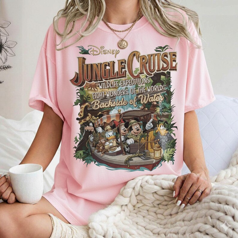 May include: A light pink t-shirt featuring a Disney Jungle Cruise graphic. The graphic includes the text "Jungle Cruise" and images of Mickey Mouse, Donald Duck, and other characters on a boat. The shirt has short sleeves.