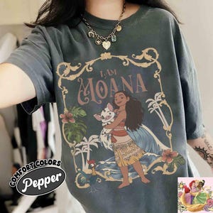 May include: Dark gray t-shirt featuring a graphic of Moana holding a pig, with the text "I AM MOANA." The design includes tropical flowers, palm trees, and a rooster. The shirt has the Comfort Colors Pepper logo.
