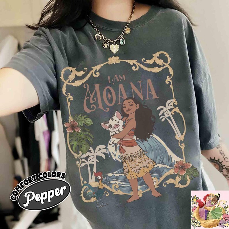 May include: Dark gray t-shirt featuring a graphic of Moana holding a pig, with the text "I AM MOANA." The design includes tropical flowers, palm trees, and a rooster. The shirt has the Comfort Colors Pepper logo.