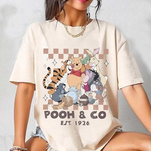 Puede incluir: Camiseta beige con un gráfico de Winnie the Pooh y sus amigos, incluyendo Tigger, Piglet, Eeyore y Rabbit. El diseño incluye el texto "POOH & CO EST. 1926" en fuente marrón, con un fondo a cuadros.