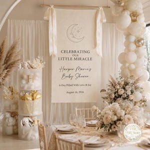 May include: A cream-colored fabric banner hanging from a wooden rod with white ribbons, featuring a crescent moon illustration and the text: CELEBRATING OUR LITTLE MIRACLE, Harper Marie's Baby Shower, A Day Filled With Love & Joy, August 16, 2026.