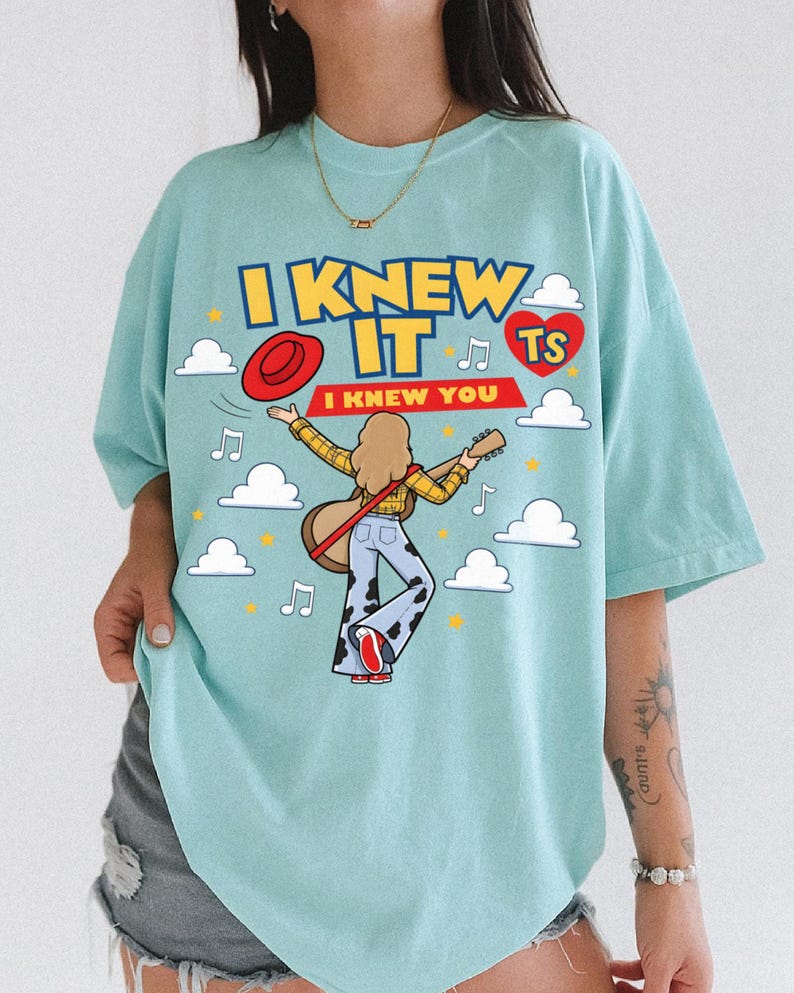 Puede incluir: Una camiseta azul claro con el gr&aacute;fico de una persona de pelo largo tocando una guitarra ac&uacute;stica, vistiendo una camisa de cuadros amarilla y pantalones con estampado de vaca. El texto dice I KNEW IT y I KNEW YOU, junto a un sombrero rojo.