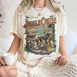 May include: Cream-colored t-shirt featuring a vintage-style Disney Jungle Cruise graphic. The design includes the text "Jungle Cruise," "Wildlife Expeditions," and "Eight Wonders of the World Backside of Water," along with cartoon characters in a boat.