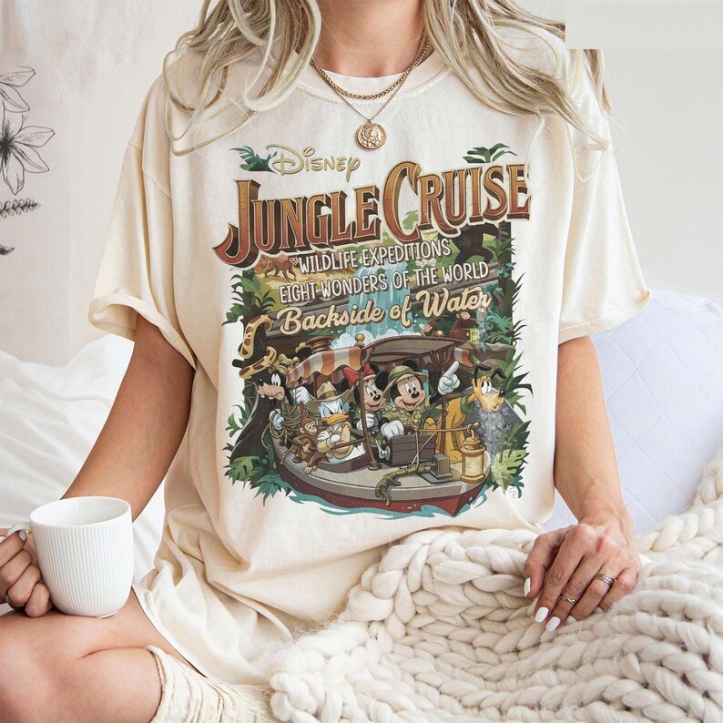 May include: Cream-colored t-shirt featuring a vintage-style Disney Jungle Cruise graphic. The design includes the text "Jungle Cruise," "Wildlife Expeditions," and "Eight Wonders of the World Backside of Water," along with cartoon characters in a boat.