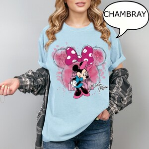 Op de afbeelding: Lichtblauw T-shirt met een Minnie Mouse-ontwerp. Het ontwerp bevat een roze strik met witte stippen, een cartoonafbeelding van Minnie Mouse en de woorden "Minnie Mouse". Het woord "CHAMBRAY" staat in een tekstballon.