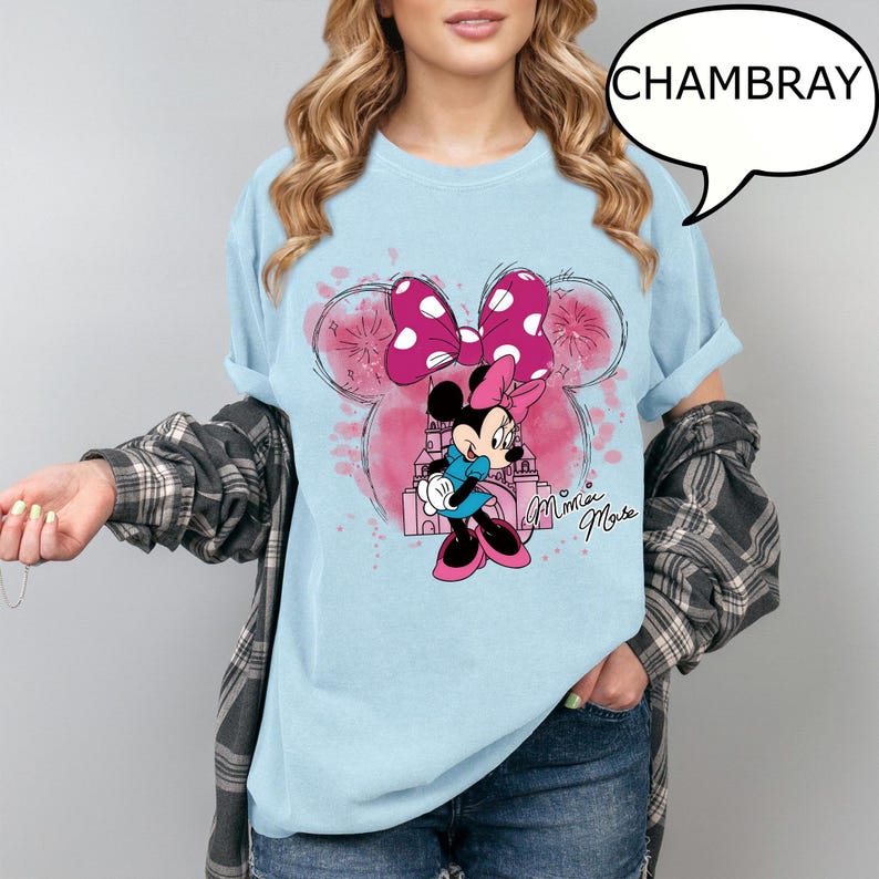 Op de afbeelding: Lichtblauw T-shirt met een Minnie Mouse-ontwerp. Het ontwerp bevat een roze strik met witte stippen, een cartoonafbeelding van Minnie Mouse en de woorden "Minnie Mouse". Het woord "CHAMBRAY" staat in een tekstballon.