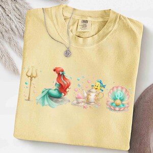 K&ouml;nnte beinhalten: Hellgelbes T-Shirt mit einem farbenfrohen Aufdruck einer Meerjungfrau mit roten Haaren und einem t&uuml;rkisfarbenen Schwanz, sowie einem goldenen Dreizack. Eine silberne Halskette mit Anh&auml;nger liegt auf dem T-Shirt.