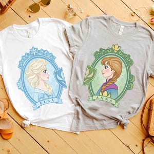 May include: Two t-shirts featuring cartoon portraits of Elsa and Anna from Frozen. The white t-shirt has a blue and white Elsa design, while the gray t-shirt has a green and yellow Anna design. Both shirts have the character's name on a banner.