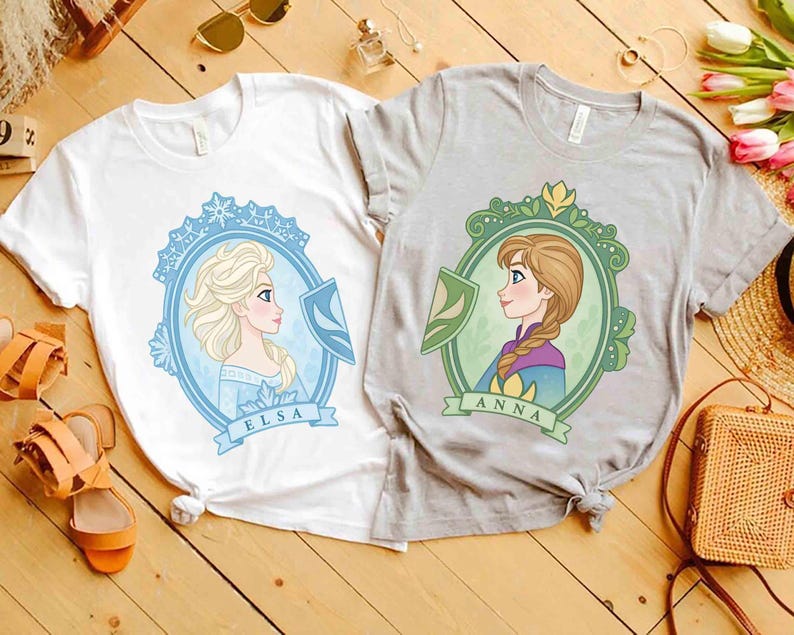 May include: Two t-shirts featuring cartoon portraits of Elsa and Anna from Frozen. The white t-shirt has a blue and white Elsa design, while the gray t-shirt has a green and yellow Anna design. Both shirts have the character's name on a banner.