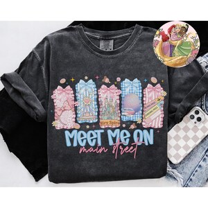 May include: Dark gray t-shirt with a colorful graphic design. The design features pink, blue, and white bows with various theme park icons and the text "Meet Me On Main Street". A cartoon of three princesses is in the upper right corner.