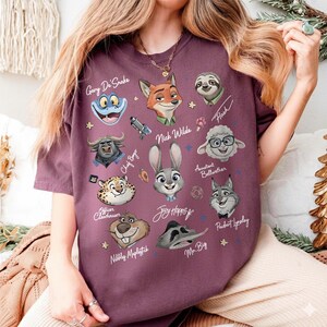 K&ouml;nnte beinhalten: Ein weinrotes T-Shirt mit Cartoon-Illustrationen von Charakteren aus einem Film. Zu den Charakteren geh&ouml;ren ein Fuchs, ein Hase, ein Faultier und andere, jeweils mit ihren Namen dar&uuml;ber oder darunter geschrieben. Das T-Shirt hat einen Rundhalsausschnitt.