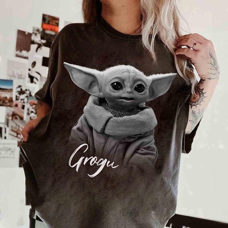 May include: Dark brown t-shirt featuring a grayscale image of Grogu, with large ears and a hooded cloak. The word "Grogu" is written in white cursive script below the image. The shirt is a casual style.