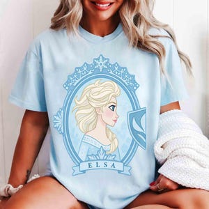 May include: Light blue t-shirt featuring a portrait of Elsa from Frozen. The design is framed in a light blue oval with snowflake accents and the name "ELSA" on a banner below. The shirt is a short-sleeved crew neck.
