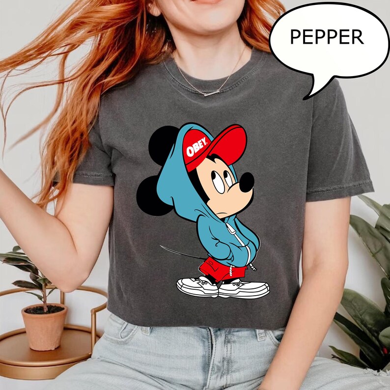 May include: Dark gray t-shirt featuring a cartoon Mickey Mouse character wearing a red cap and blue hoodie. The word "OBEY" is printed on the cap. The word "PEPPER" is in a speech bubble.