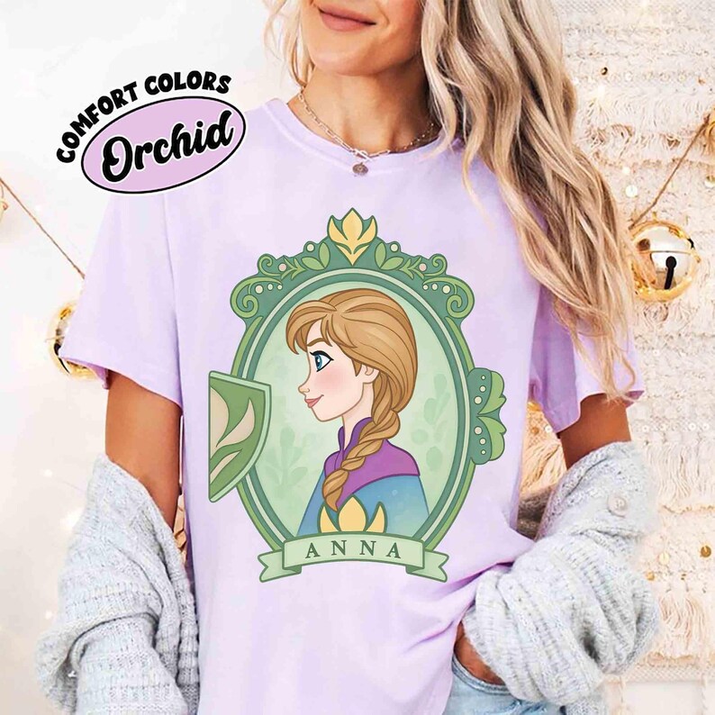 May include: A light purple t-shirt with the text "COMFORT COLORS Orchid" at the top. The shirt features a cartoon illustration of a woman with a braid, inside a green and gold frame, with the name "ANNA" below.