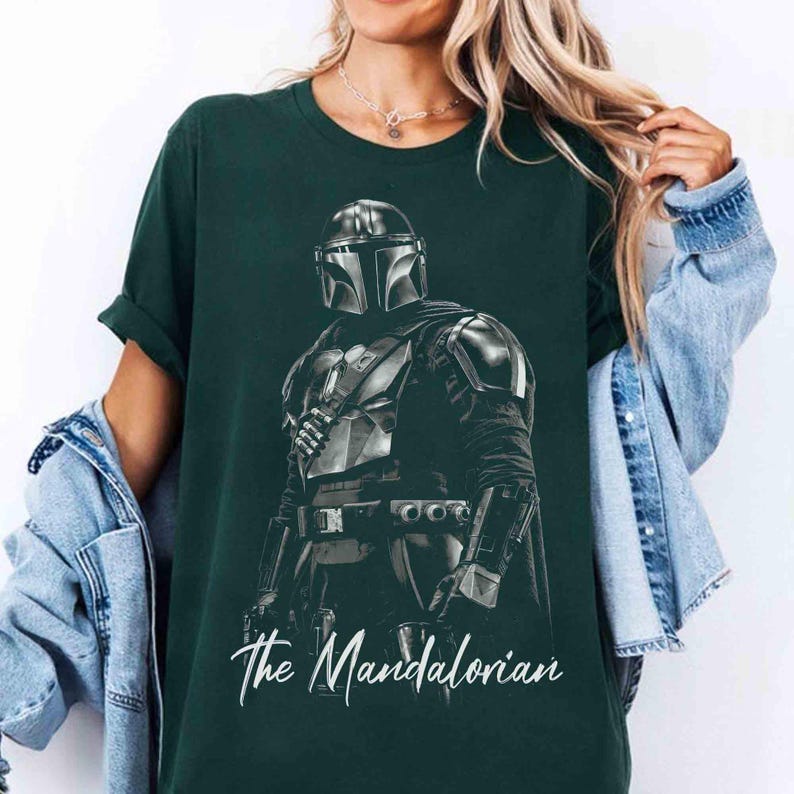 May include: Dark green t-shirt featuring a detailed black and white illustration of The Mandalorian. The graphic depicts the character in full armor, with the text "The Mandalorian" in a cursive font at the bottom.