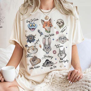 K&ouml;nnte beinhalten: Cremefarbenes T-Shirt mit Illustrationen von Zeichentrickfiguren aus Zootopia, darunter Nick Wilde, Judy Hopps und Flash. Das Shirt hat Charakternamen und Sternakzente. Das Shirt ist kurz&auml;rmelig.