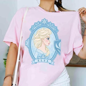 May include: A light pink t-shirt featuring a portrait of Elsa from Frozen. The portrait is framed in a light blue oval with snowflake and crown details. The name "ELSA" is printed on a banner below the portrait.