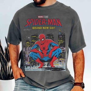 K&ouml;nnte beinhalten: Dunkelgraues T-Shirt mit einer Grafik von Spider-Man in einer hockenden Pose vor einer Stadtkulisse. Das Shirt zeigt den Text "MARVEL STUDIOS SPIDER-MAN BRAND NEW DAY" in roter und blauer Schrift.