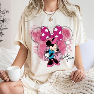 Op de afbeelding: Cr&egrave;mekleurig T-shirt met een Minnie Mouse-afbeelding. Het ontwerp bevat een roze strik met witte stippen, een cartoonafbeelding van Minnie Mouse en de woorden "Minnie Mouse" in cursief. De achtergrond heeft roze aquareleffecten.