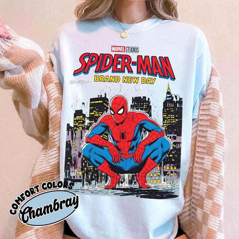 K&ouml;nnte beinhalten: Hellblaues Comfort Colors Chambray Sweatshirt mit einem Marvel Studios Spider-Man-Grafik. Das Design zeigt die Worte "Spider-Man Brand New Day" und eine Illustration von Spider-Man, der sich vor einer Skyline hockt.