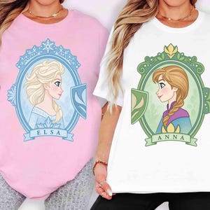 Könnte beinhalten: Zwei T-Shirts, eines rosa und eines weißen, jeweils mit einem Cartoon-Porträt einer Prinzessin. Das rosa Shirt zeigt Elsas Porträt, während das weiße Shirt Annas Porträt zeigt. Beide Porträts sind mit dekorativen Elementen und Namen umrahmt.