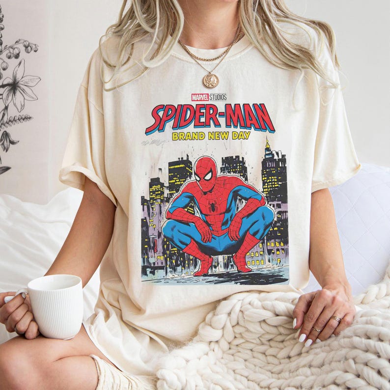 K&ouml;nnte beinhalten: Cremefarbenes T-Shirt mit einer Grafik von Spider-Man in einer hockenden Pose vor einer Stadtkulisse. Der Text "MARVEL STUDIOS SPIDER-MAN BRAND NEW DAY" ist in Rot und Blau &uuml;ber dem Bild gedruckt.