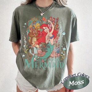 May include: A moss green short-sleeve t-shirt featuring a graphic print of Ariel from The Little Mermaid with Flounder, surrounded by underwater elements like bubbles, seaweed, and coral. The text THE LITTLE MERMAID is printed in teal below.