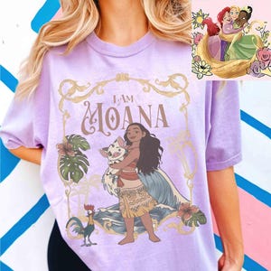 May include: A light purple t-shirt featuring the phrase "I AM MOANA" in a decorative font. The graphic includes an illustration of Moana holding a pig, with a rooster and tropical flowers. A smaller image of Disney princesses is in the upper right corner.