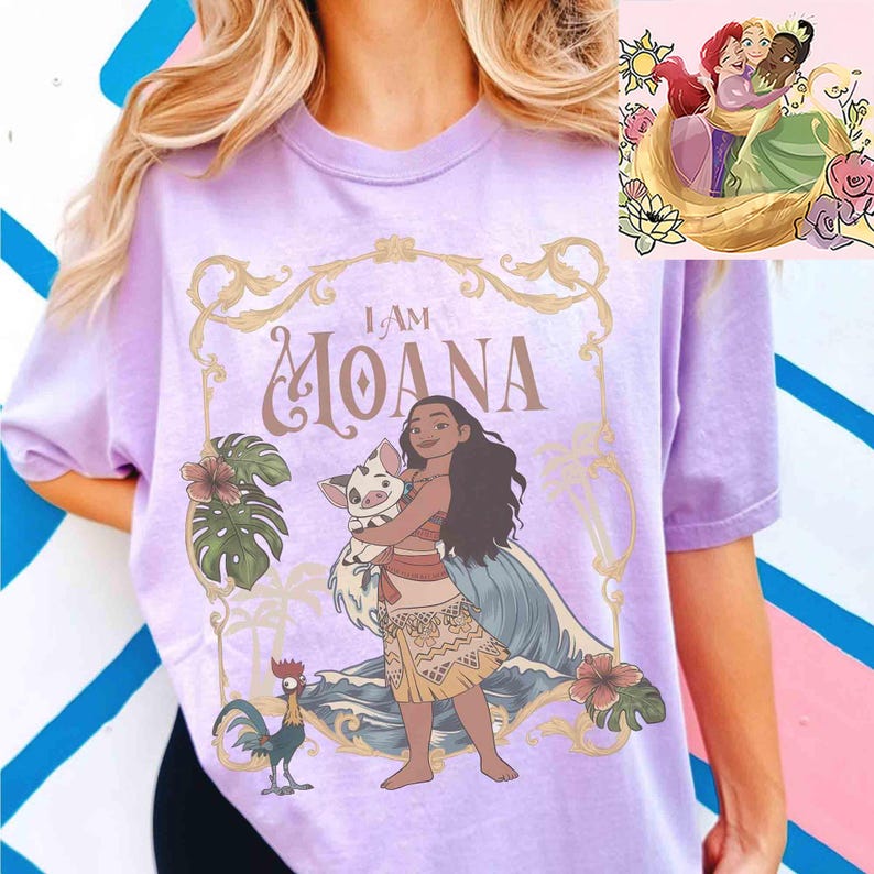 May include: A light purple t-shirt featuring the phrase "I AM MOANA" in a decorative font. The graphic includes an illustration of Moana holding a pig, with a rooster and tropical flowers. A smaller image of Disney princesses is in the upper right corner.