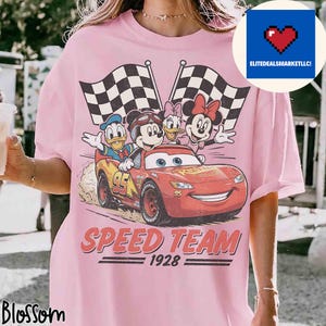 Pu&ograve; includere: T-shirt rosa chiaro con una grafica dei personaggi Disney Topolino, Paperino, Paperina e Minnie in un'auto da corsa rossa con bandiere a scacchi. Il testo "SPEED TEAM 1928" &egrave; stampato sotto.