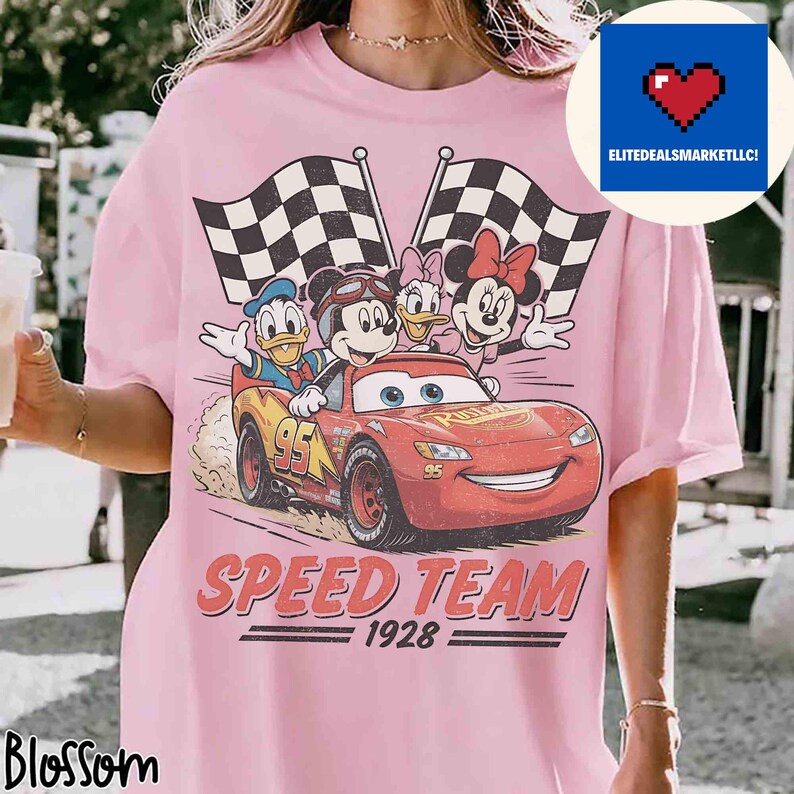 Pu&ograve; includere: T-shirt rosa chiaro con una grafica dei personaggi Disney Topolino, Paperino, Paperina e Minnie in un'auto da corsa rossa con bandiere a scacchi. Il testo "SPEED TEAM 1928" &egrave; stampato sotto.