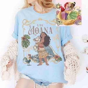 May include: Light blue t-shirt featuring a graphic of Moana holding a pig, with the text "I AM MOANA." The design includes floral and wave elements. A smaller image of three princesses is in the upper right corner. The shirt has rolled-up sleeves.